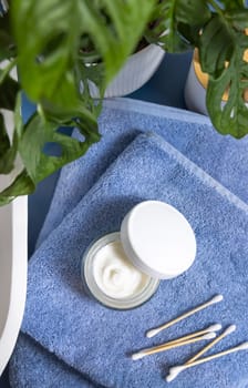 Opened cream jar with blank white lid and bamboo cotton swabs on blue towels near basin and green monstera blue countertop, top view, mockup. Lifestile scene with skincare beauty product in bathroom