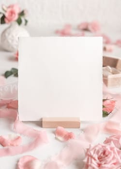 Blank square card near light pink roses, petals, engagement ring in a box and silk ribbons on white table close up, wedding mockup. Romantic scene with envelope and pastel decor