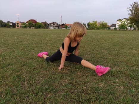 A 6-7 year old girl goes in for sports at the stadium. A child in sportswear in black and bright pink sneakers. Exercise for stretching the muscles of the legs Split. Caucasian girl with blond hair.