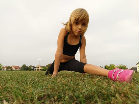 A 6-7 year old girl goes in for sports at the stadium. A child in sportswear in black and bright pink sneakers. Exercise for stretching the muscles of the legs Split. Caucasian girl with blond hair.