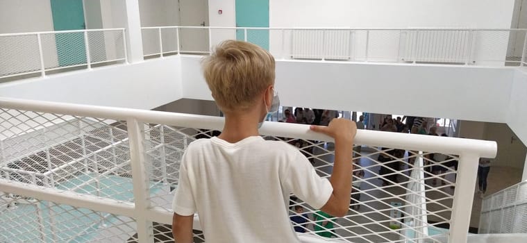 Sremska Mitrovica, Serbia, September 1, 2020 A boy in a white shirt and mask stands near a metal fence. A child of 7 years old holds onto the handrail. A modern school or clinic with white walls.