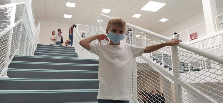 Sremska Mitrovica, Serbia, September 1, 2020. A boy in a white shirt and a mask poses on the stairs. A child of 7 years old holds onto the handrail. Modern school, clinic or hospital with white walls.