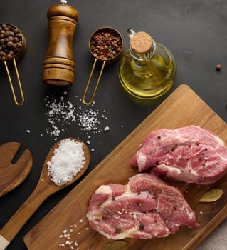 Two raw pork neck steaks on a board and spices for cooking. Top view of black table.