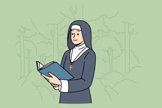 Woman nun with holy book stands in park among trees, dressed in cassock for religious service in temple. Nun studies sacred scriptures and prayers during breaks between prayer services in cathedral