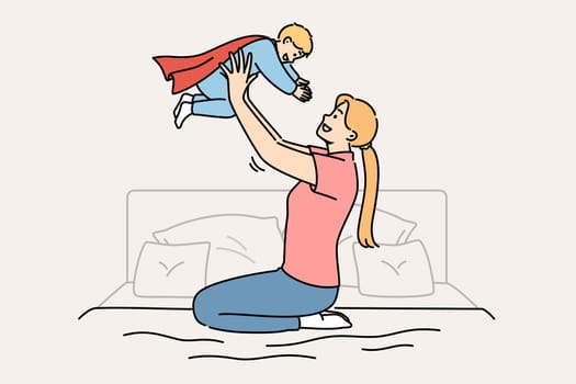 Happy mother lifts up newborn son in superhero costume, sitting on bed in bedroom. Caring casual mother plays with child, motivating son to achieve ambitious goals and success in life