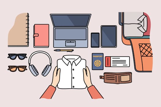 Personal items for business travel and suitcase for luggage are laid out on table near passport for customs control. Hands of man preparing clothes and gadgets for flight on business trip
