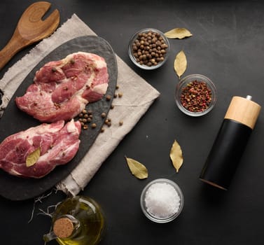 Two raw pork neck steaks on a board and spices for cooking. Top view of black table
