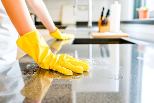 Hands in yellow gloves cleaning a dark countertop with a sponge, sparkling clean surface reflected.