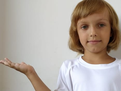 A little cute girl 6-7 years old with blond hair expresses emotional joy and a smile on a white background. A child with tanned skin. White T-shirt. Copy space. Girl with her hand points to something.