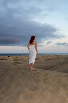 A young girl stands in a white dress in the sand dunes with sky and clouds in the late afternoon