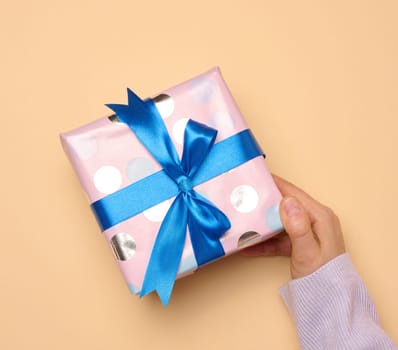 A woman's hand holds a gift box wrapped in a blue silk ribbon on a beige background, top view