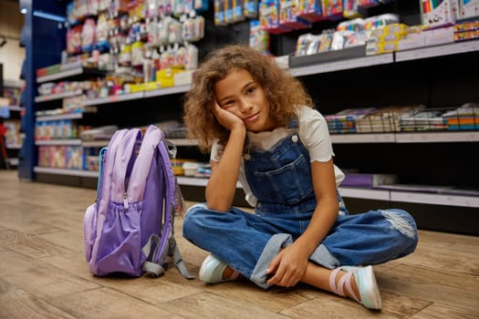 Bored or tired girl child sitting on floor of stationery shop. Cute little female student feeling exhausted after shopping to prepare for studying