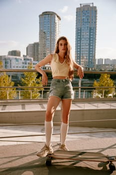 Cool hipster woman skateboarder posing with her longboard over cityscape full-length portrait. Beautiful sportive teenager girl enjoying training at downtown district. Youth culture and sport concept