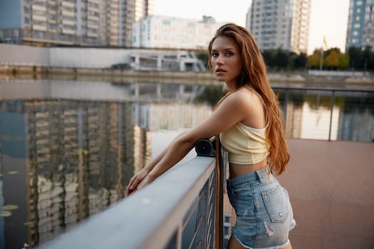 Charming attractive girl wearing casual clothes having relax time standing with longboard nearby city river. Portrait of young teenage woman with perfect body, sensual look holding skateboard