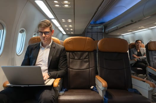 Handsome businessman with eyeglasses working on laptop computer while sitting in airplane cabin. Internet connection on board service and business travel concept.