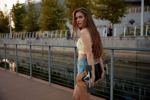 Sensual slim young woman with tanned skin wearing casual clothes walking with longboard over summer cityscape