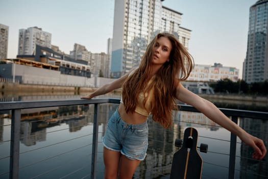 Young teenage woman enjoying time for skateboarding through city street. Beautiful female posing for camera standing with longboard over urban river in city park