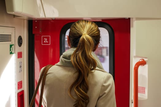 Young woman in coat standing next to stopping train door, about to leave - view from behind