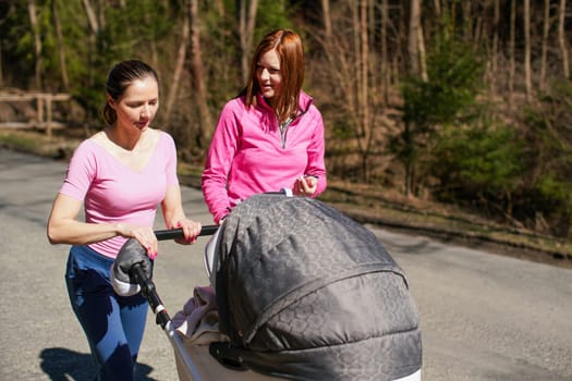 Two young woman in pink tops walking with baby carriage over asphalt road trees on side, sunny day