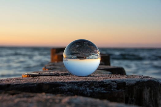 Glass ball lies on the wood of a breakwater on the beach at sunset, the sea and the setting sun are reflected in the ball