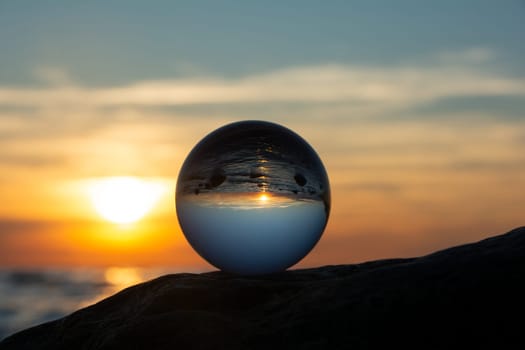 Glass ball on a rock at sunset on the beach, the sea and the setting sun are reflected in the ball