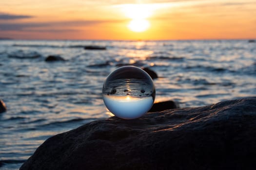 Glass ball on a rock at sunset on the beach, the sea and the setting sun are reflected in the ball