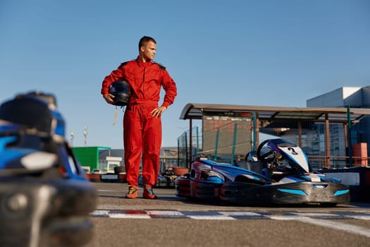 Equipped go-kart driver wearing overalls and holding helmet walking to car before race at starting line on motor racing track. Automotive sports activity, carting championship