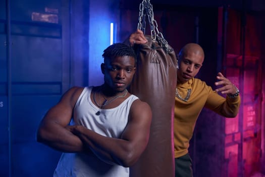Young gang boys training with punching bag in garage. Two hipster guys friends exercising pumping muscles together