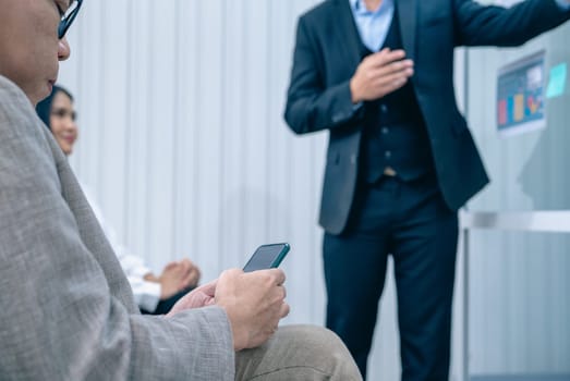A man is standing in front of a group of people and pointing to a screen. A woman is sitting in the background and looking at her phone. The man is giving a presentation