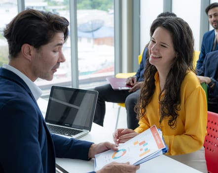 A man and a woman are sitting at a table with a laptop and a stack of papers. The woman is smiling and she is interested in the presentation