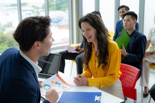 A group of people are sitting around a table with a woman in a yellow shirt. The woman is smiling and talking to a man. The man is looking at a laptop. Scene is friendly and collaborative
