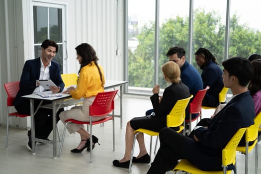 A group of people are sitting in chairs in a room, some of them are wearing suits. They are all looking at a man who is reading something. Scene is professional and serious