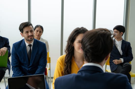A group of people are sitting in a room, some of them wearing suits. A man in a suit is talking to a woman in a yellow dress. Scene is professional and formal