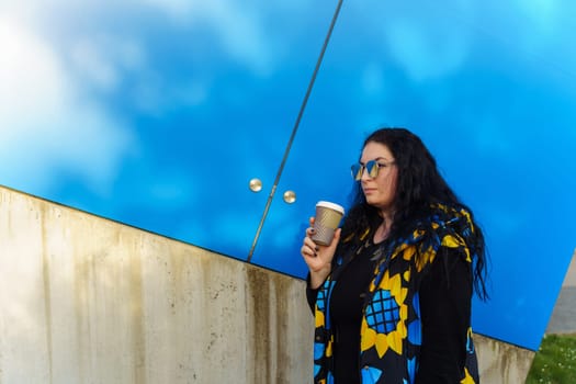 Beautiful young brunette woman with long curly hair, dressed in a black, blue and yellow waistcoat, drinks coffee from a paper cup on a background of blue wall.