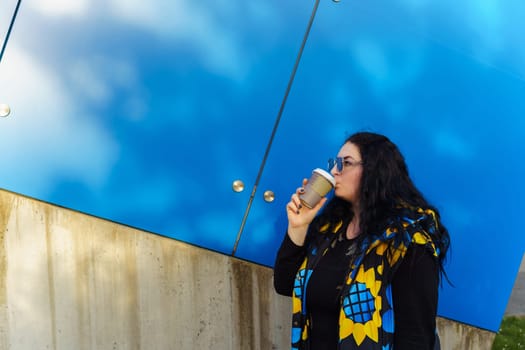 Beautiful young brunette woman with long curly hair, dressed in a black, blue and yellow waistcoat, drinks coffee from a paper cup on a background of blue wall.