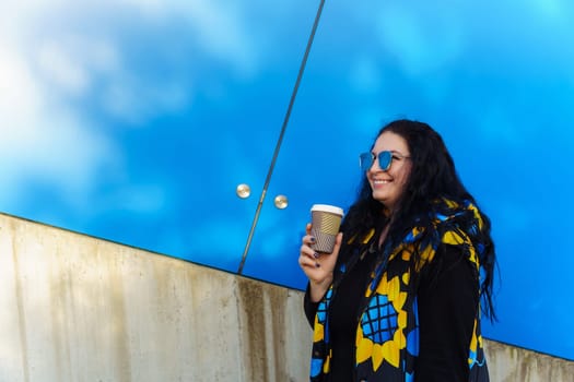 Beautiful young brunette woman with long curly hair, dressed in a black, blue and yellow waistcoat, drinks coffee from a paper cup on a background of blue wall.