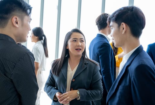 A group of people are standing in a room, some of them wearing suits. A woman is talking to another person, and they are both smiling. Scene is friendly and professional