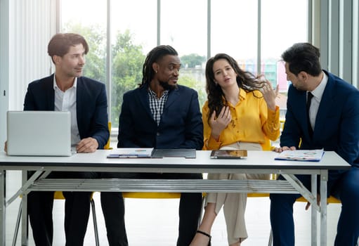 A group of people are sitting around a table in a business setting. One woman is talking to the group while the others listen. The atmosphere is professional and serious