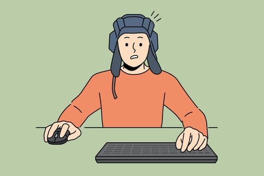 Man gamer plays tanks on computer, sitting at table with keyboard and wears tanker helmet. Guy gamer passionate about video games and virtual tank simulators, makes embarrassed grimace after losing