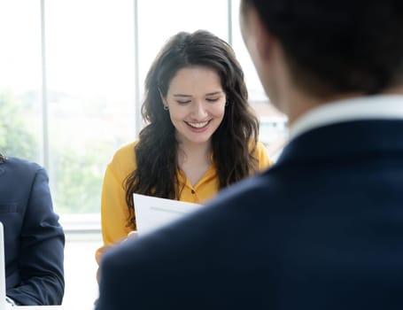 A woman in a yellow shirt is reading a piece of paper while a man in a suit watches her. The scene appears to be a business meeting or a professional setting
