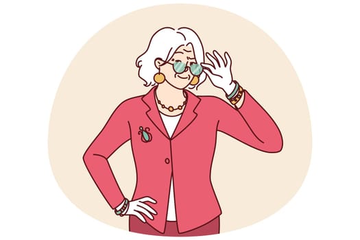 Elderly woman in elegant suit for going to work or dinner party adjusts glasses in front of eyes. Gray-haired grandmother of retirement age dressed in fashionable elegant clothes looks at camera
