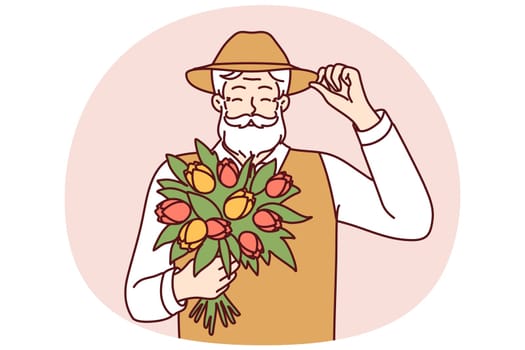 Cheerful elderly man holds bouquet of spring flowers touches hat in greeting for happy old age concept. Positive gray-haired man with mustache and beard holds flowers tulips grown in own garden