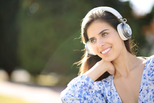 Happy woman looks away listening audio on headphone sitting in a garden