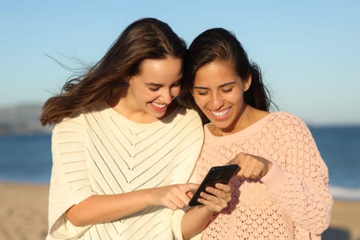 Two happy friends talking about phone content on the beach