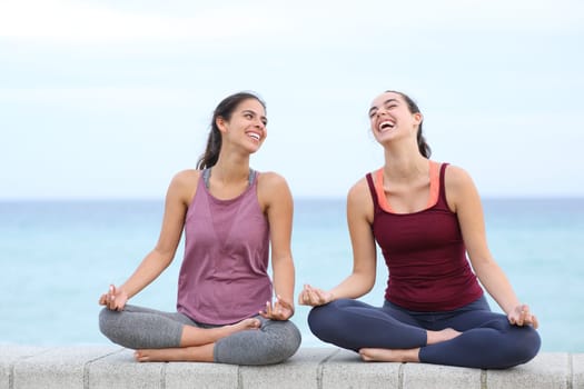 Front view portrait of two happy women doing yoga laughing on the beach