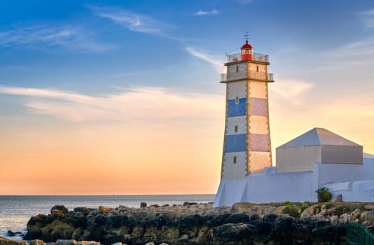 Beautiful sunset on sea shore and Santa Marta lighthouse in Cascais, Portugal. Colorful sky, clouds, sunlight, low sun, calm ocean waters, local landmark, safety and navigation tower, rocky shore.