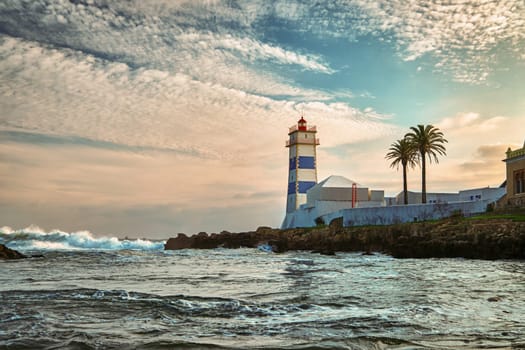 Beautiful sunset landscape on sea shore and Santa Marta lighthouse in Cascais, Portugal. Colorful sky, clouds, sunlight, low sun, calm ocean waters, local landmark, safety and navigation tower, rocky shore.