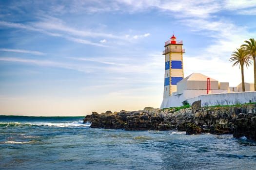 Beautiful evening, on sea tidal waves, rocky shores and Santa Marta lighthouse in Cascais, Portugal. Blue sky, clouds, sunlight, low sun, calm ocean waters, local landmark, safety and navigation tower, palm trees.