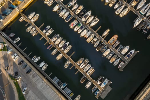 Aerial top down view of a row of yachts, sailboats and jetties in marina with moored sailboats. Low sun, sunshine, drone stay and ascend revealing more vessels.