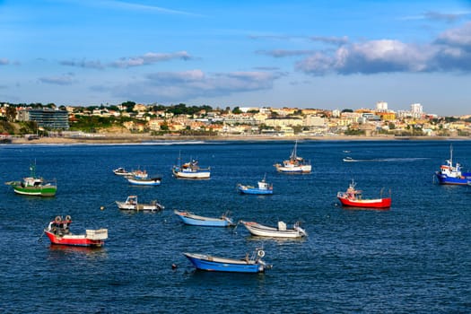 Beautiful view of small fishing boats on sunny day in bay of Cascais, Portugal, quiet and luxurious seaside town near Lisbon with beaches and small port. Clear blue sky, clouds, sunshine, calm ocean waters, houses on coastline.